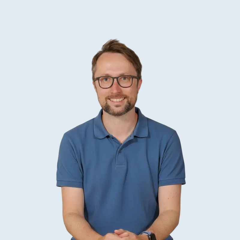 Mann im blauen Polohemd sitzt lächelnd auf einem Stuhl vor hellem Hintergrund.